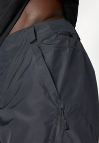 Dunkelgraue wasserdichte Hose mit samtweicher Textur, schrägen Taschen und einer mit Reißverschluss versehenen Seitentasche für sicheren Stauraum.