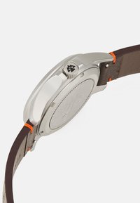 Boîtier de montre en acier inoxydable argenté avec un bracelet en cuir brun. Détail de couture orange et couronne texturée ajoutent un intérêt visuel.