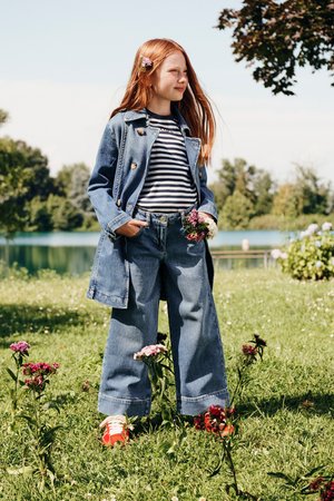 Jeansjacke mit Messingknöpfen, gestreiftem Hemd, weitem Denim-Jeans und roten Schuhen. Haare mit kleinen Blumen geschmückt. Gras und Blumen im Hintergrund.