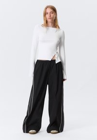Weißes Langarmshirt mit seitlichen Knopfdetails, kombiniert mit lockeren schwarzen Hosen mit weißen Seitenstreifen und einer strukturierten Stoffoberfläche.