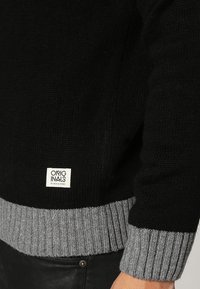 Pull en maille noir avec un ourlet gris côtelé, présentant une étiquette tissée en bas avec le nom de la marque. Gros plan sur la manche.