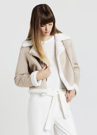 Giacca in finta suede beige con collo e polsini in pelliccia bianca, chiusura con zip, design cropped, indossata sopra una maglietta bianca e pantaloni a vita alta.