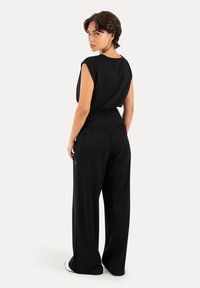 Femme portant un haut noir sans manches et un pantalon noir à jambes larges, debout, regardant par-dessus son épaule sur un fond clair uni.