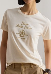Γυναίκα με εκρού μπλουζάκι Lauren Ralph Lauren με χρυσό κεντημένο λογότυπο και καφέ παντελόνι, στέκεται μπροστά σε απλό φόντο.