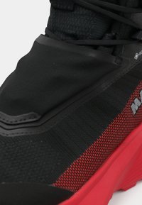 Zwarte en rode sportieve schoen met een gestructureerde bovenkant, gladde overlays en een uitgesproken rode middenzool met subtiele logo's aan de zijkant.