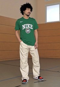 Grøn Nike t-shirt med hvidt logo, beige cargo bukser og sorte/røde sneakers. Baggrunden har vægge af træ og et basketballgulv.