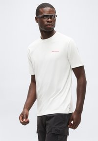 Άνδρας που φοράει λευκό T-shirt Helly Hansen, μαύρα cargo σορτς και αθλητικά σκούρα γυαλιά ηλίου, στέκεται μπροστά σε απλό φωτεινό φόντο.