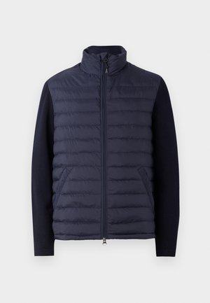 Navy puffer jacket with a high collar, zip front, and padded body. Knitted sleeves and side pockets create a textured contrast.