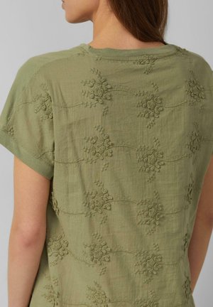 Versleten olijfgroene top met korte mouwen en geborduurde bloemenpatronen op de rug, zichtbaar van schouders tot middenrug.