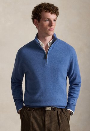 Jeune homme aux cheveux bouclés portant un pull bleu à fermeture quart de zip sur une chemise blanche et un pantalon marron à carreaux, les mains dans les poches.