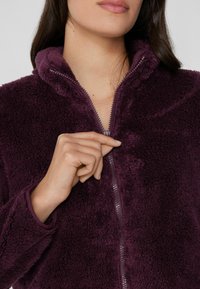 Chaqueta de forro polar morado oscuro con cremallera frontal. Presenta un cuello alto y una tela texturizada. Primer plano de una mano sosteniendo la cremallera.
