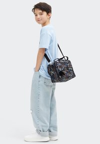 Borsa a tracolla nera con motivi colorati a tema skateboard, materiale testurizzato, doppia cerniera e spallacci regolabili, indossata da una persona in abbigliamento azzurro chiaro.