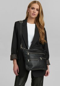Borsa a tracolla nera in pelle con tre tasche con zip, hardware silver, e una texture morbida, indossata sopra una camicia bianca e un blazer nero.