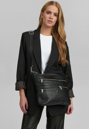 Schwarze Leder-Crossbody-Tasche mit drei Reißverschlussfächern, silbernem Zubehör und weicher Textur, getragen über einem weißen Hemd und einem schwarzen Blazer.