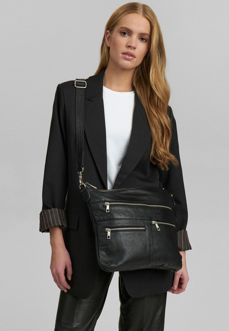 Borsa a tracolla nera in pelle con tre tasche con zip, hardware silver, e una texture morbida, indossata sopra una camicia bianca e un blazer nero.