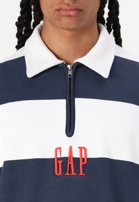Polo a rayas en azul marino y blanco con cuello con cremallera, que presenta un logo bordado en rojo "GAP" en la parte frontal. Tejido suave con una textura lisa.