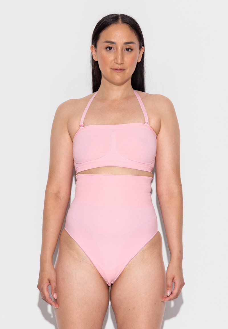 Frau mit langen dunklen Haaren, die einen hellrosa High-Waist-Bikini mit einem trägerlosen Oberteil und abnehmbaren Neckholder-Trägern trägt, vor einem weißen Hintergrund steht.