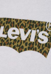 T-shirt grafico di Levi's con uno sfondo in tessuto bianco che presenta un logo leopardato verde e marrone. Texture liscia, lettering audace.
