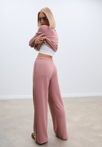 Ružičasti set za opuštanje koji se sastoji od krojenog topa i hlača širokih nogavica s elastičnom gumicom. Glatka tekstura i opušten silueta.