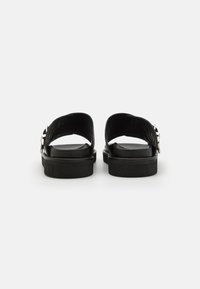 Sandale din piele neagră cu talpă plată, spate descoperit și detaliu cu cataramă. Partea superioară are un design elegant cu o textură netedă.