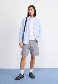 Camisa de botones a rayas azules y blancas, pantalones cortos grises, zapatillas negras con textura y calcetines blancos. Conjunto informal con detalle de bolso bandolera.