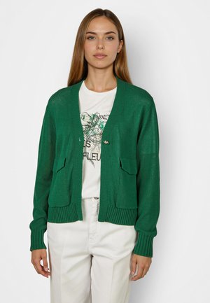 Woman wearing green buttoned cardigan over white graphic t-shirt and white pants, standing against a plain light background.