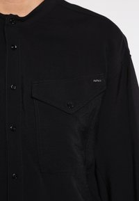 Camisa negra de botón, hecha de una tela suave. Cuenta con un bolsillo en el pecho con un botón a presión y un detalle de logo sutil. Brillo leve en el material.