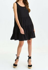 Schwarzes ärmelloses Kleid mit quadratischem Ausschnitt, gestuften Rüschen und fließender Silhouette, kombiniert mit schwarzen Espadrilles.