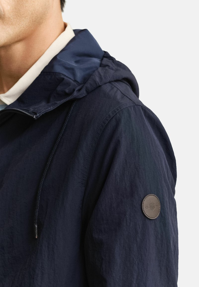 Homme portant une veste à capuche bleu marine avec des cordons de serrage et une petite écusson rond en cuir sur le haut de la manche, sur un fond clair.