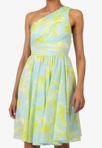 Robe à épaule asymétrique en tissu bleu clair avec des motifs floraux jaunes ; le corps plissé et la jupe froncée créent une silhouette fluide.