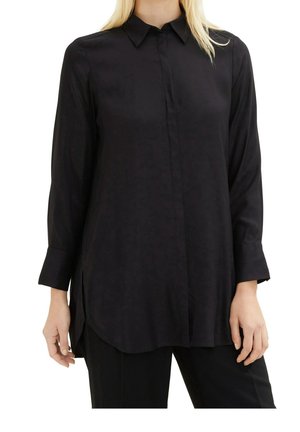 Kobieta ubrana w luźną, długą czarną koszulę na guziki z kołnierzykiem i długimi rękawami, zestawioną z czarnymi spodniami.