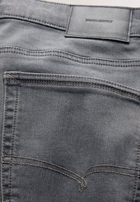 Grå denimjeans baglomme med kontrasterende brun syning og et lædermærke mærket "Denim Supply" på taljebåndet.