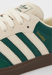 Grön och beige sneaker med tre ränder, texturerad mocka och läder, med "SAMBA" i guld på sidan och en brun gummisula.
