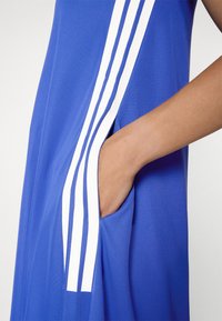 Blue athletic dress in textured fabric, featuring white vertical stripes and a side pocket. Close-up on the design and pocket detail.