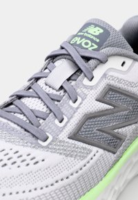 Chaussure de sport légère en mailles grises et blanches, dotée d'une semelle à épaisse couche de mousse verte, d'accents réfléchissants et de lacets plats gris.