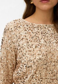Top de lentejuelas beige con cuello redondo, mangas largas y una textura brillante formada por lentejuelas de varios tamaños en tonos dorados y bronces.
