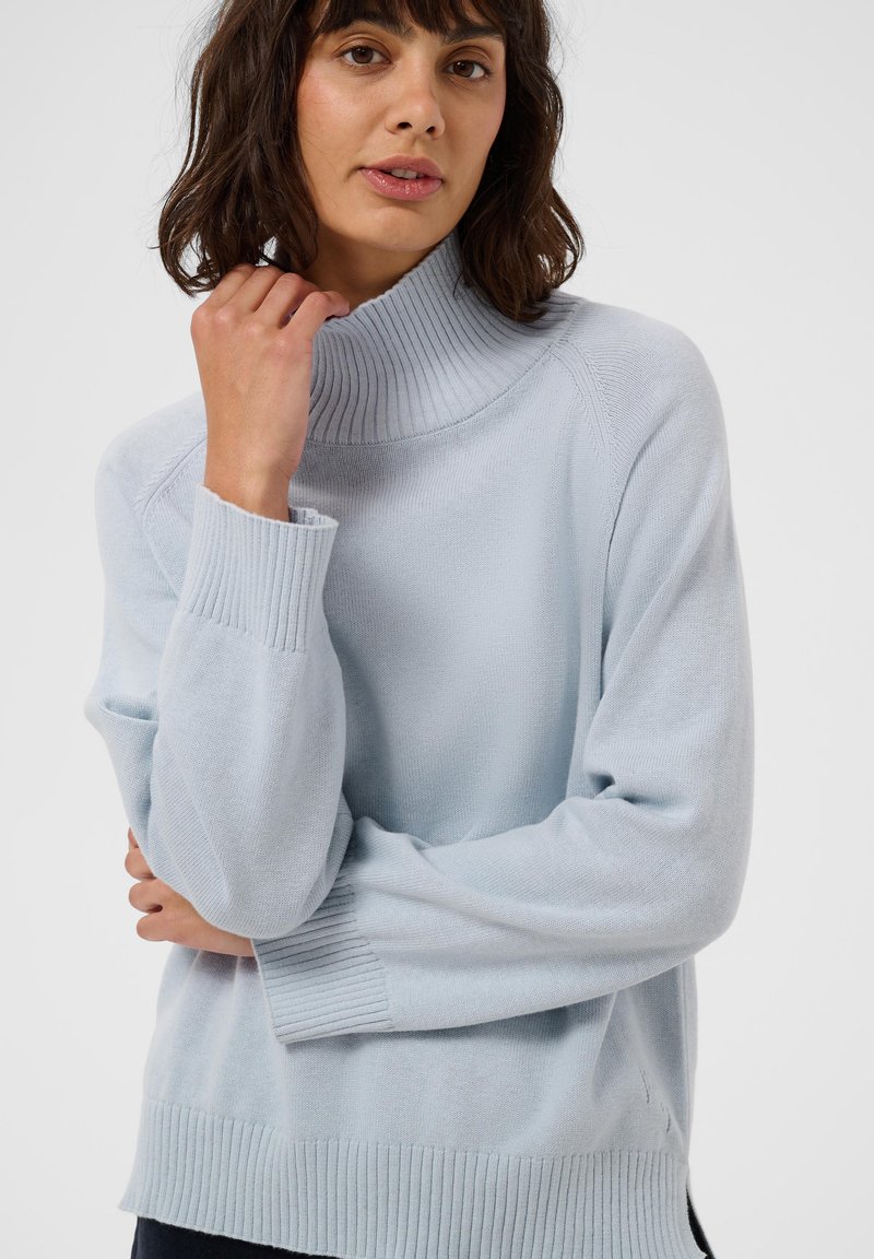 Femme aux cheveux foncés portant un pull à col roulé côtelé bleu clair, tenant le col d'une main et croisant les bras.