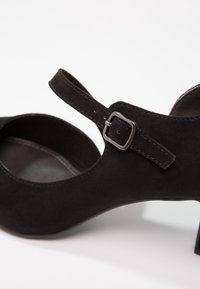 Chaussure à talons hauts en daim noir avec design à bout ouvert, bride de cheville avec boucle argentée, texture lisse et talon légèrement incurvé.