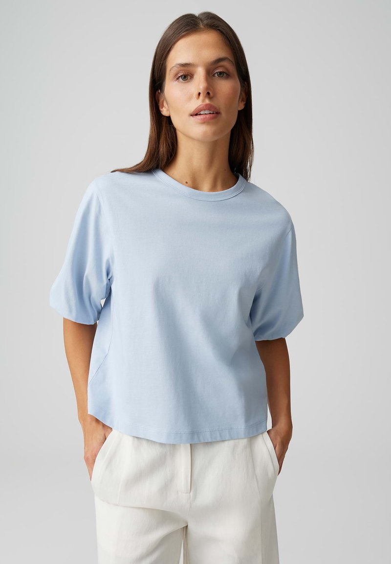 Junge Frau mit glatten braunen Haaren, die ein hellblaues kurzärmeliges Hemd und weiße Hose trägt, Hände in den Taschen, neutraler Hintergrund.