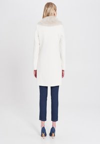 Cappotto bianco con colletto in pelliccia sintetica, maniche lunghe, taglio dritto, abbinato a jeans slim blu scuro e tacchi, visto da dietro.