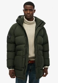 Ciemnozielona kurtka puchowa z kapturem, z dużymi pikowanymi sekcjami, bocznymi kieszeniami i czarnym detalem naszywki. Noszona na kremowym golfie.