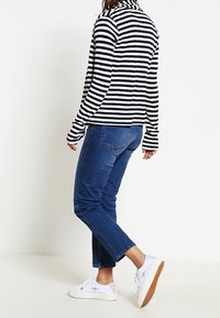 Persona vestida con una camiseta de rayas en blanco y negro de manga larga, jeans recortados azules y zapatillas blancas con suelas marrones, de pie de lado.