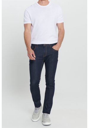 Homme portant une chemise blanche à manches courtes, un jean slim bleu marine et des baskets gris clair, se tenant avec une main dans la poche.