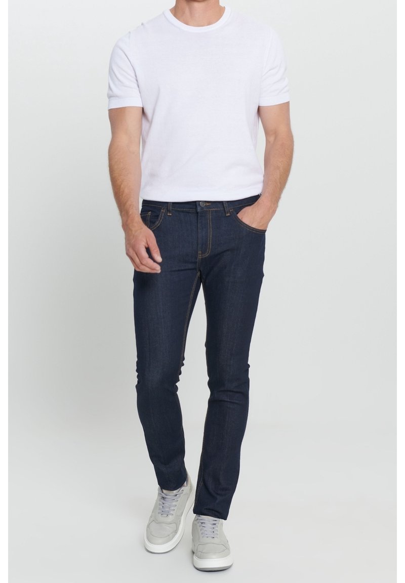 Homme portant une chemise blanche à manches courtes, un jean slim bleu marine et des baskets gris clair, se tenant avec une main dans la poche.