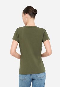 T-shirt verde oliva a maniche corte con scollo rotondo, texture liscia e silhouette aderente, mostrato da dietro con jeans di denim.
