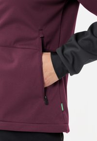 Bordeauxrote Jacke mit glatter Textur, ausgestattet mit einer Reißverschlusstasche und schwarzen Ärmeln. Beinhaltet ein kleines grünes Etikett am Saum.