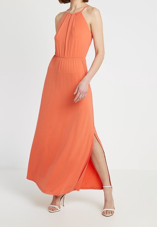 Femme portant une robe maxi orange sans manches avec une fente latérale et des sandales blanches à talons hauts, debout devant un fond blanc uni.