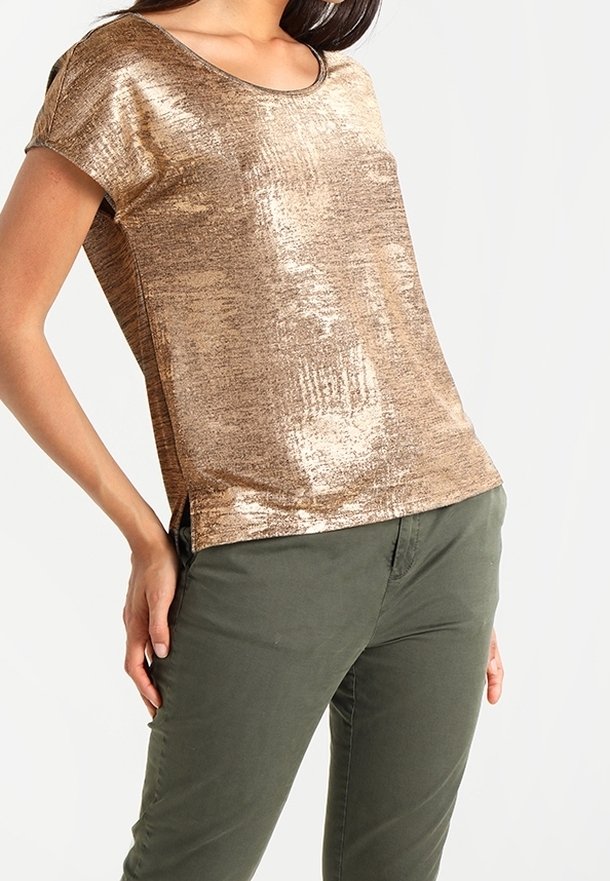 Kobieta ubrana w błyszczącą złotą bluzkę z krótkim rękawem, zestawioną z oliwkowozielonymi spodniami, stojąca z rękami przy biodrach na jednolitym tle.