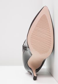 Scarpa con tacco alto in vernice nera, con punta affusolata e tacco sottile. La suola è beige con scanalature testurizzate per migliorare la presa.