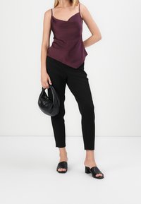 Camisole en satin bordeaux avec un ourlet diagonal, associé à un pantalon noir taillé et des sandales à glisser noires. Le modèle tient un sac hobo noir.